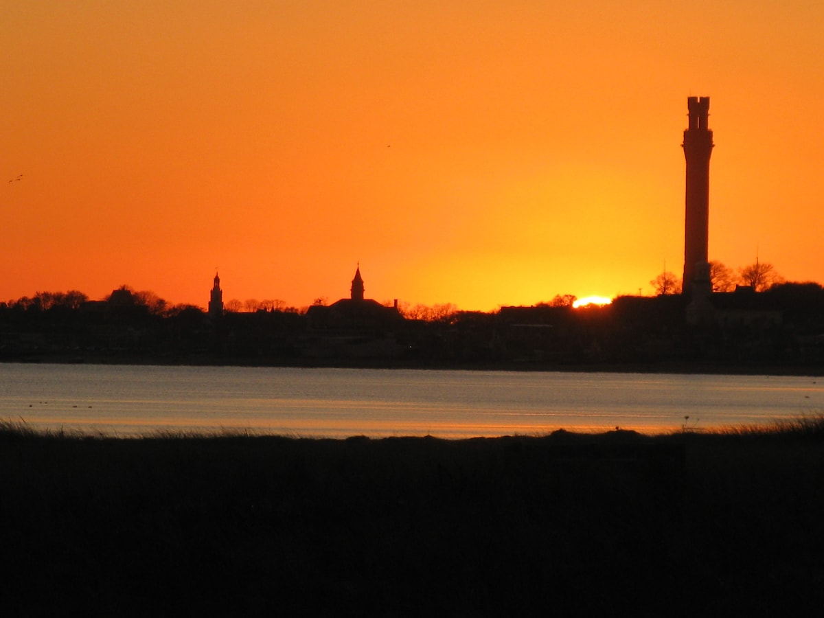 A dramatic sunset illuminates the horizon with shades of orange and yellow, silhouetting the distant buildings and monuments along the shore. The calm water reflects the warm colors, creating a serene atmosphere over the landscape.