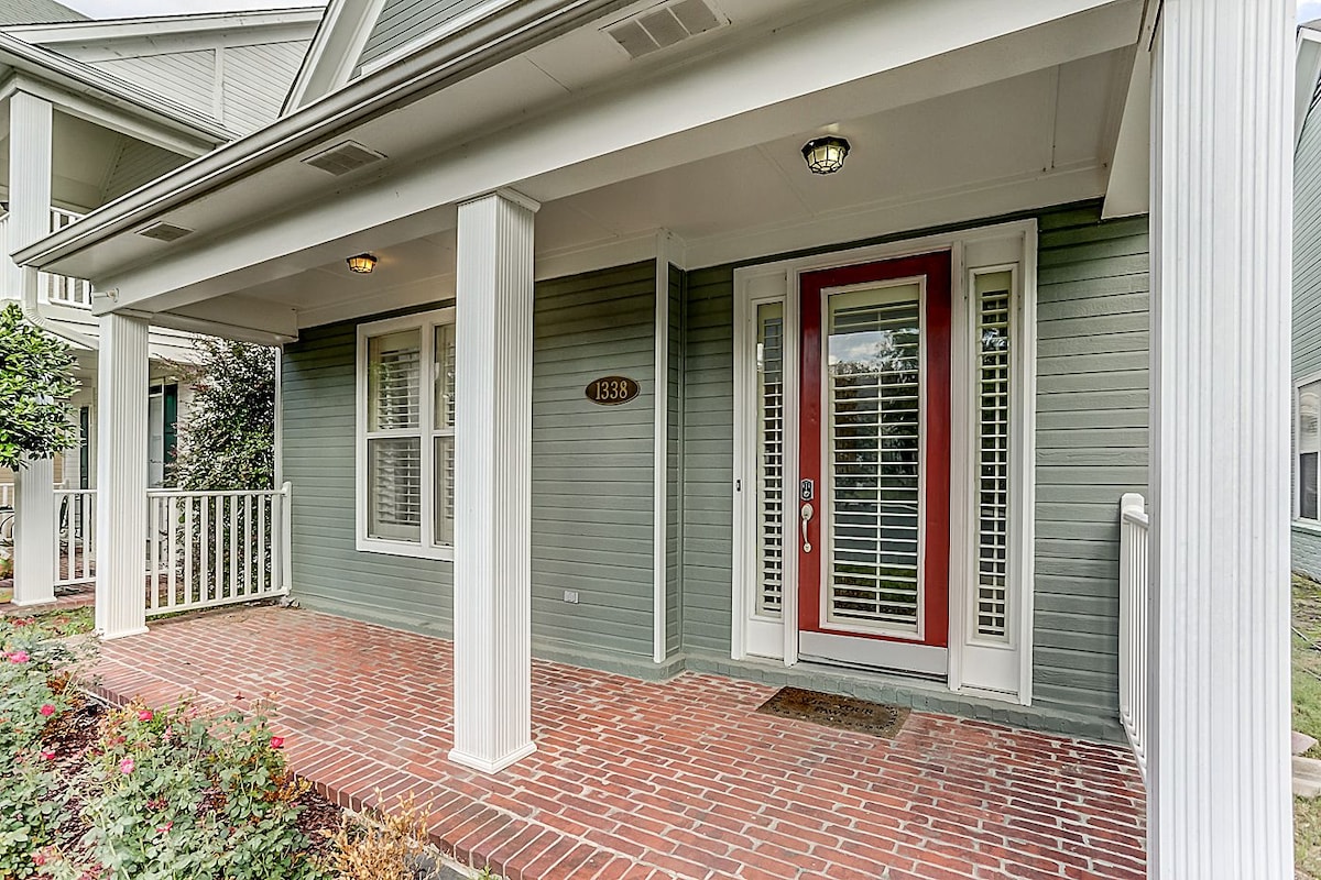 The entrance to the home features a welcoming covered porch with brick flooring. White columns provide structural support, while a red front door offers a focal point. Windows with plantation shutters contribute to privacy, and symmetrical landscaping adds a touch of greenery.