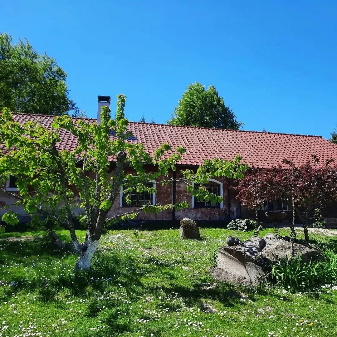 A charming cottage with a red tiled roof is surrounded by a lush green lawn. Trees with fresh foliage and flowering plants add color to the landscape. The clear blue sky complements the peaceful outdoor setting.