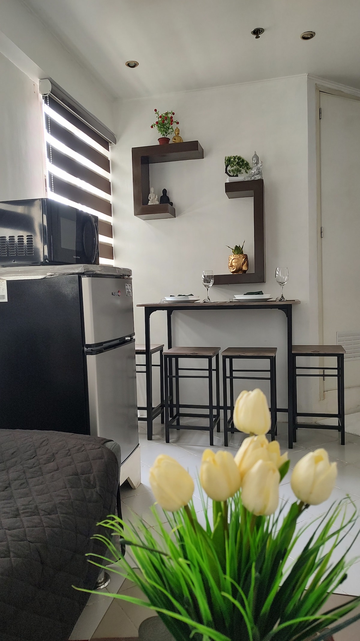A compact dining area features a small table set for two, complemented by sleek black stools. A mini refrigerator stands nearby, and decorative shelves showcase potted plants and figurines. Fresh flowers in a planter add a touch of greenery to the space, enhancing the overall ambiance.