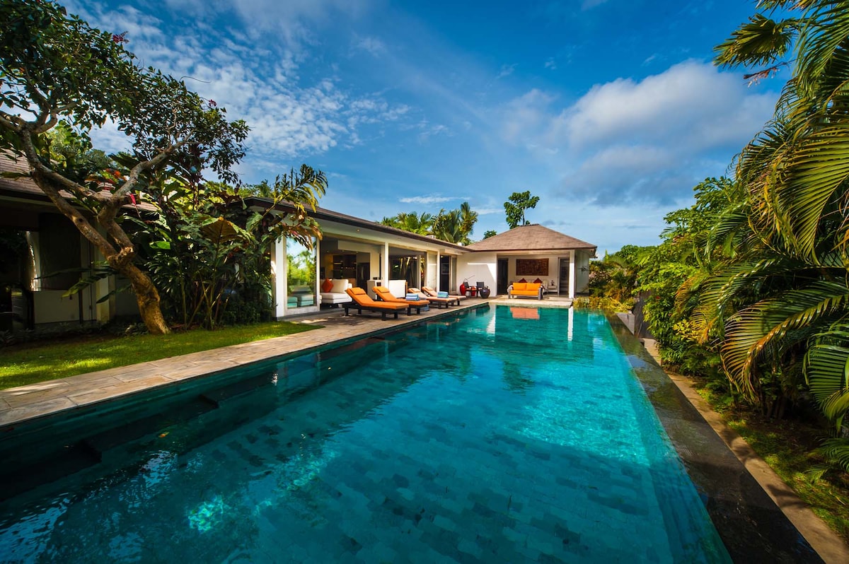 A private pool stretches invitingly in the foreground, flanked by sun loungers for relaxation. Behind, the villa's open design is visible, blending harmoniously with the surrounding greenery and vibrant tropical landscape under a clear blue sky.