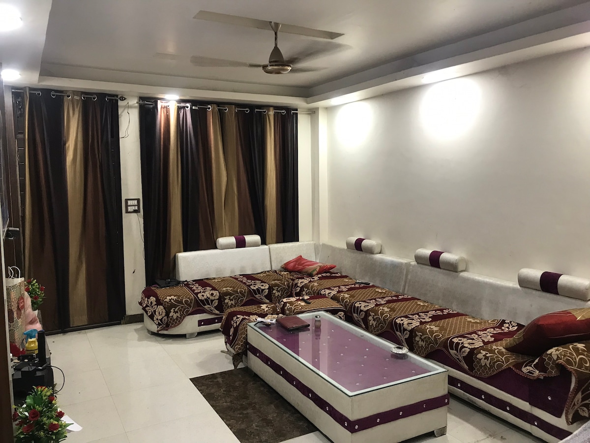 A spacious living room is shown, featuring a large seating area with cushioned benches adorned with decorative throws. The floor is tiled in white, and a glass-top coffee table is positioned at the center. Dark curtains frame the large windows, complementing the room's neutral tones.