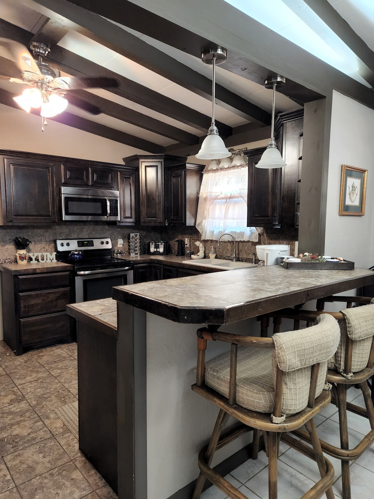 A functional kitchen showcases dark wood cabinetry and modern stainless steel appliances. The bar area features two high-backed stools, and light pours in through a window adorned with a sheer curtain. Beamed ceilings add character to the inviting space.