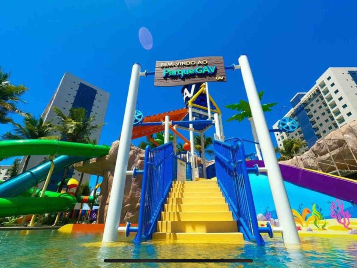 A vibrant water park entrance features colorful slides and a large staircase leading down to the water. Bright tropical-themed decorations and lush palm trees surround the area, while a clear blue sky enhances the lively atmosphere.
