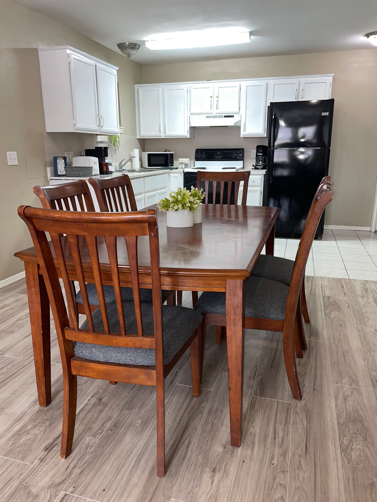 A wooden dining table is surrounded by four backrest chairs, set within a well-lit kitchen area. Light-colored cabinets contrast with a black refrigerator and kitchen appliances. A small plant decorates the table, adding a subtle touch of greenery.
