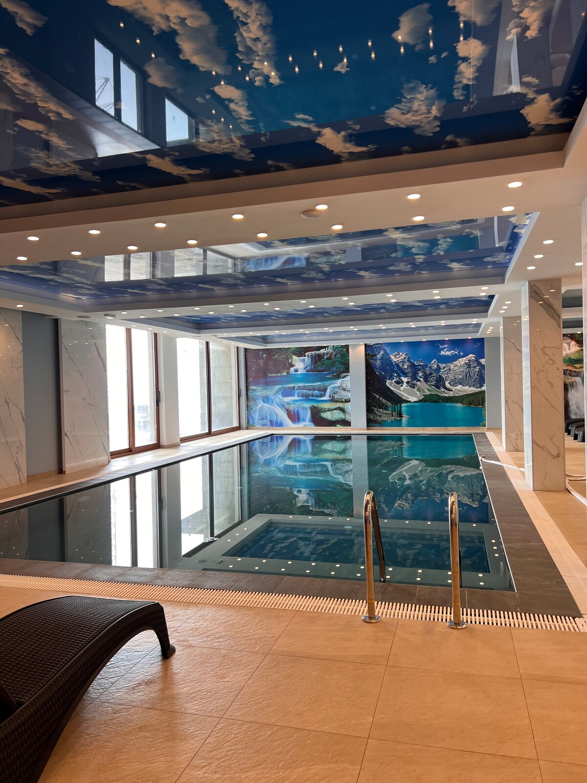 An indoor pool area is highlighted by a striking ceiling mural of clouds and mountains. The tranquil water reflects the artwork above, while large windows allow natural light to fill the space. A lounge chair is positioned nearby for relaxation.