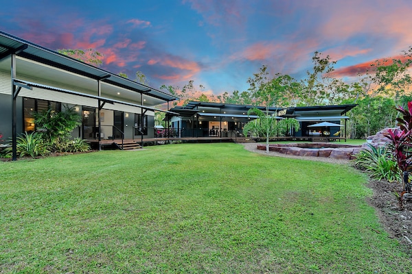 The spacious outdoor area presents a large expanse of green lawn surrounded by tropical landscaping. Multiple buildings are visible against the backdrop of a colorful sky at dusk, with deck areas providing access to each space. A peaceful atmosphere characterizes this inviting retreat.