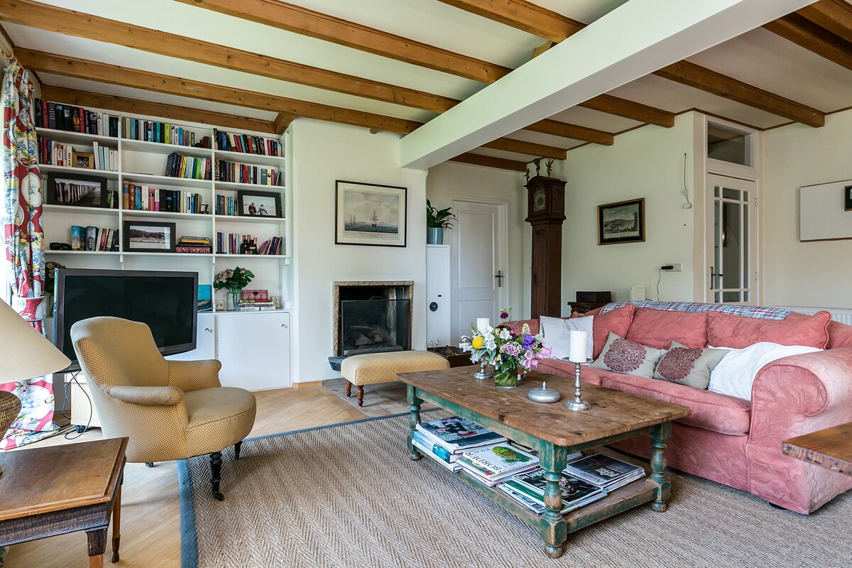 A spacious living area features wooden beams on the ceiling and a cozy seating arrangement. A pink sofa is situated next to a coffee table adorned with magazines and a bouquet of fresh flowers. A built-in bookshelf and a television are also present in the room.