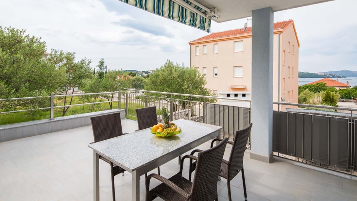 A spacious outdoor balcony is furnished with a dining table and four chairs. Natural light illuminates the area, which is surrounded by greenery and offers views of neighboring buildings and the distant coastline. A decorative fruit centerpiece is placed on the table.