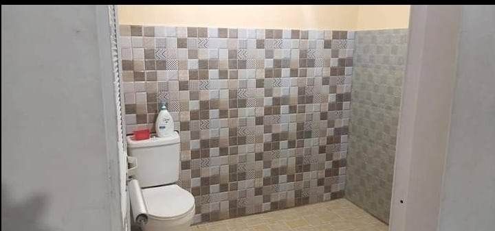 A bathroom is shown with patterned tile walls in neutral tones. A white toilet is positioned against one wall, and basic toiletries are placed on a shelf nearby. The spacious floor area features a simple design to complement the overall aesthetic.