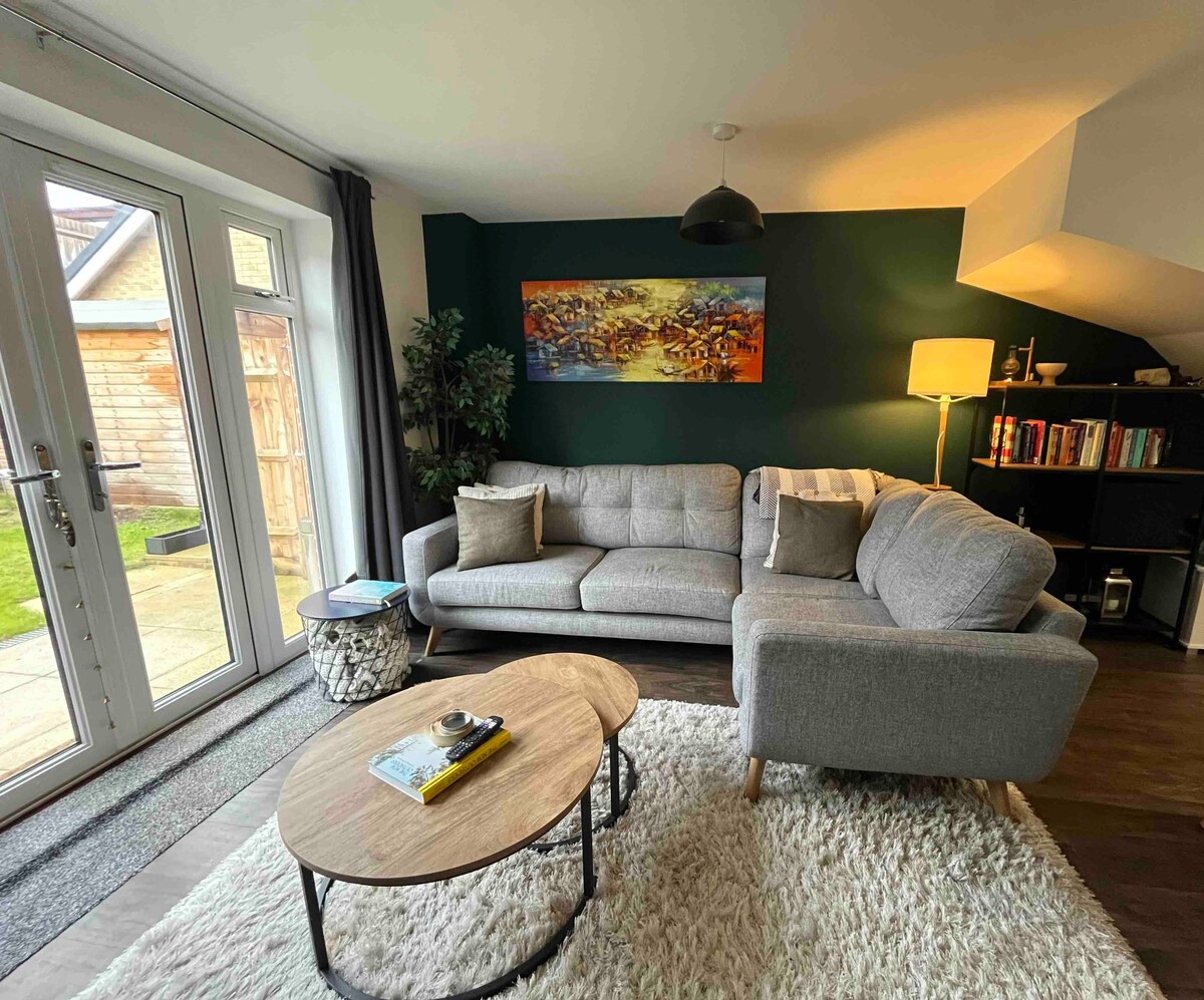 A cozy living area features a grey sectional sofa adorned with cushions, positioned beside a wooden coffee table. A statement wall in dark green is complemented by a large piece of artwork. Natural light filters through glass doors opening to a small outdoor space.