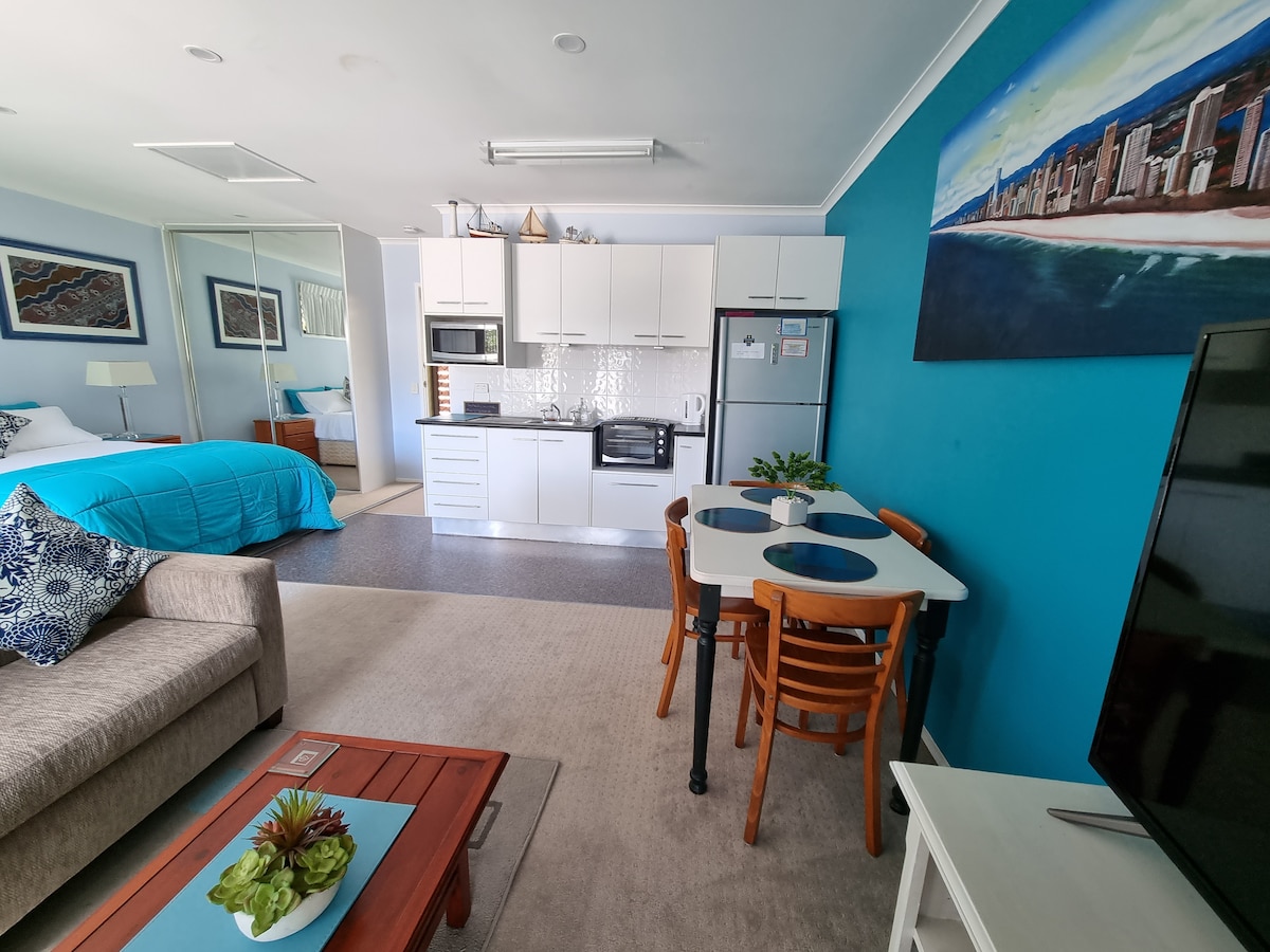 A comfortable studio space with a vibrant blue accent wall. A spacious bed features a light blue coverlet, and a well-equipped kitchenette is visible in the background. A light dining table with wooden chairs is situated beside a cozy seating area, enhancing the open layout.