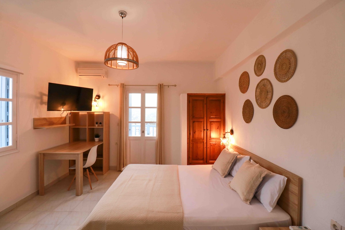 A bright room features a spacious bed with neutral bedding and two decorative pillows. A work desk with a television is positioned nearby. On the wall, circular woven art pieces add texture, while a warm light fixture hangs above, complementing natural light from the windows.