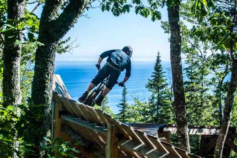 A mountain biker is captured in motion on a wooden ramp, surrounded by lush trees. The expansive view of the water is visible in the background, highlighting the natural beauty of the landscape. The scene conveys an outdoor adventure vibe.
