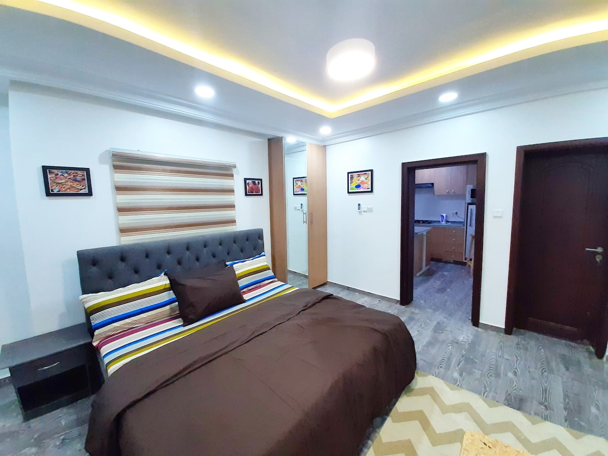 A spacious room features a king-size bed adorned with a dark brown coverlet and colorful striped pillows. Soft lighting highlights the textured walls and sleek furnishings. A doorway leads to a kitchenette, while framed artwork adds subtle interest to the serene environment.