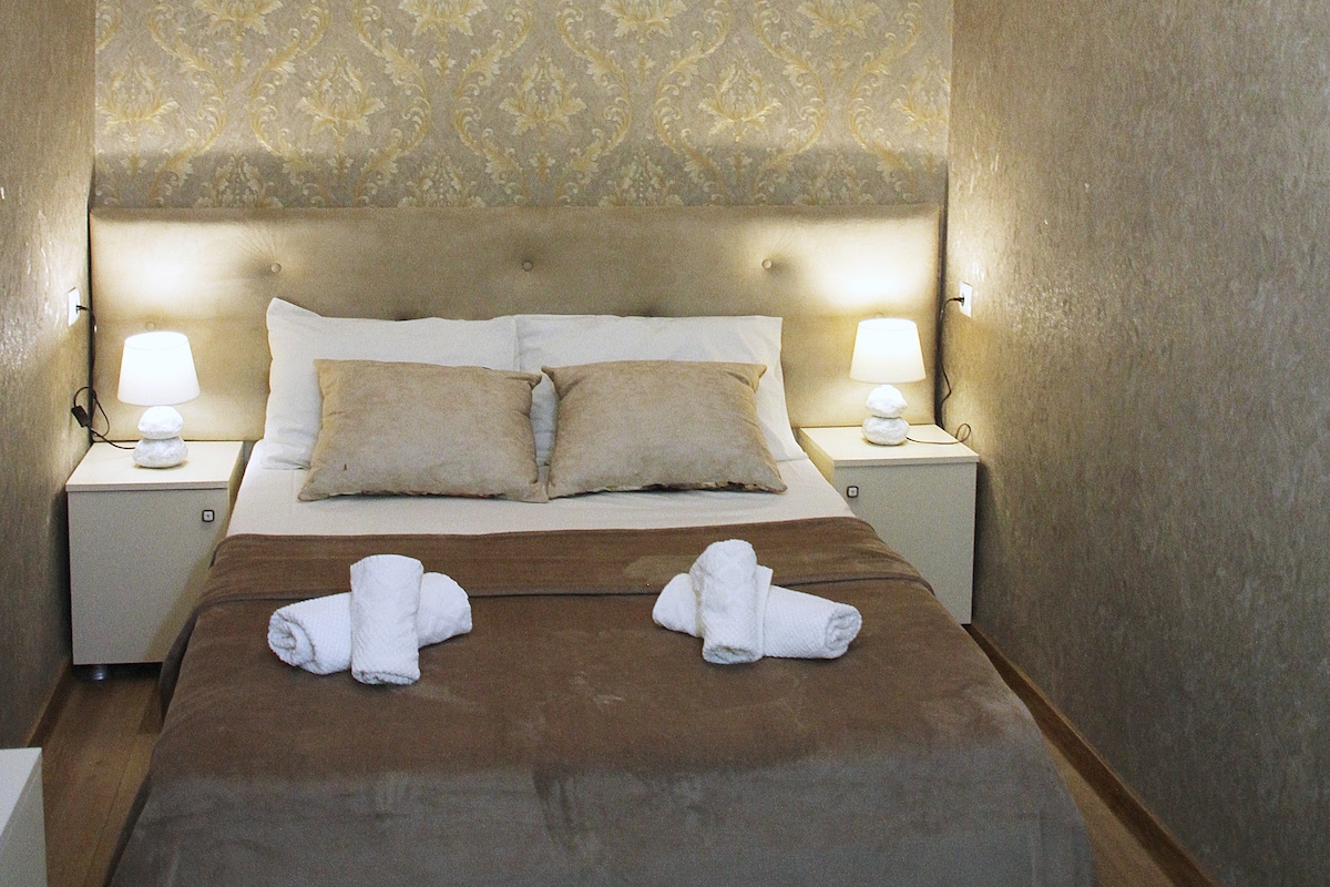 A comfortable sleeping area is highlighted with a beige upholstered headboard. Two lamps are positioned on either side of the bed, providing soft illumination. Fresh towels are neatly arranged at the foot of the spacious bed, against a backdrop of textured golden wallpaper.