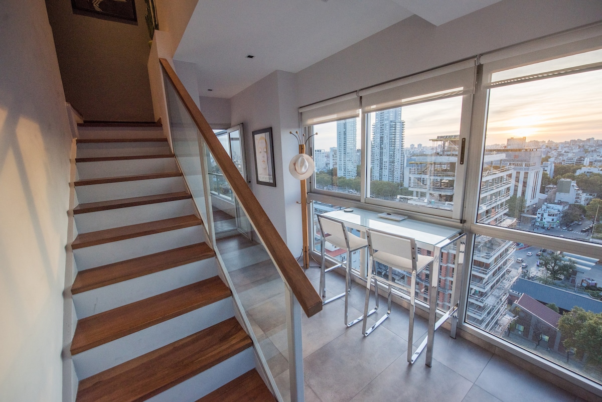 A bright stairway with wooden steps leads to an upper level. A compact workspace with two bar stools is positioned next to large windows, showcasing panoramic views of the city skyline. Soft light streams in from the setting sun, creating a warm ambiance.