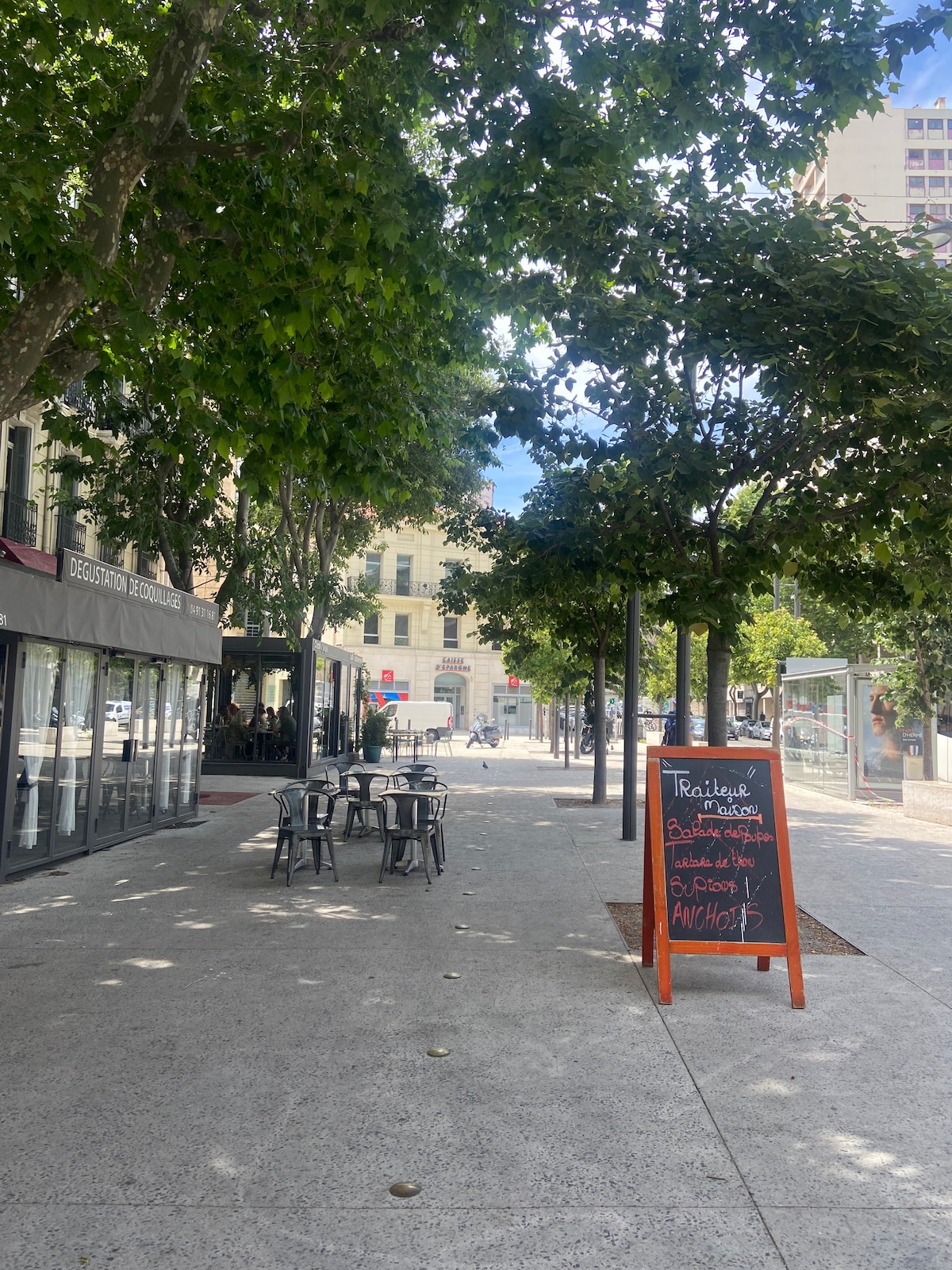 A wide, tree-lined pedestrian area is visible, featuring a mix of outdoor dining tables and a chalkboard sign promoting a restaurant nearby. The smooth concrete path is shaded by the trees, providing a calm and inviting atmosphere. Buildings are seen in the background.