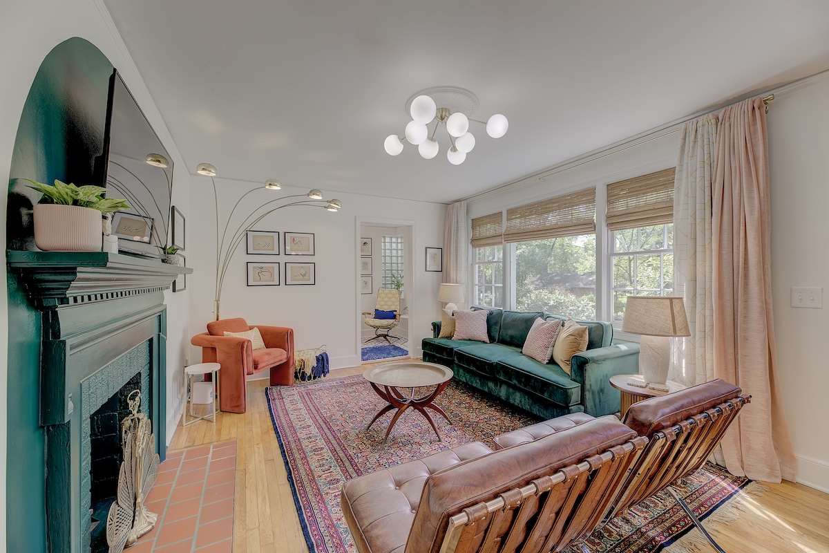 A cozy living room is presented, featuring a teal sofa, a brown leather chair, and a vintage rug. An arched floor lamp and soft lighting enhance the space. Large windows allow ample natural light, illuminating framed artwork on the walls.