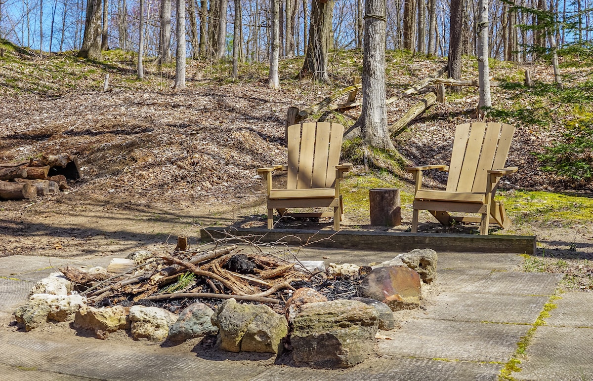 A serene fire pit area is surrounded by nature, featuring two wooden Adirondack chairs positioned beside a circular arrangement of stones and firewood. The ground is covered with a mix of soft earth and natural foliage, inviting moments of relaxation and conversation.