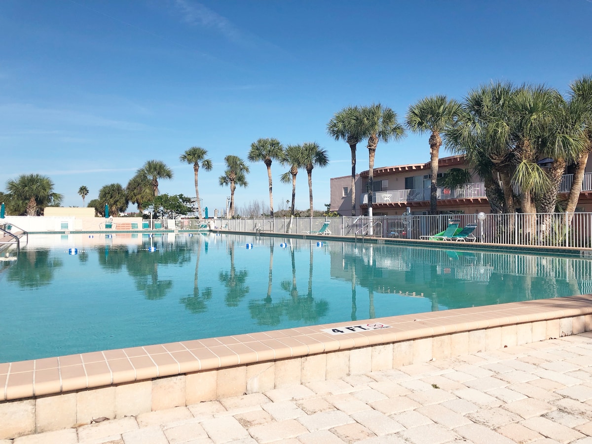A large outdoor pool is visible, surrounded by palm trees and poolside lounge chairs. The clear water reflects the blue sky, creating a serene atmosphere. The pool area is fenced and well-maintained, with sun loungers situated along the water's edge.