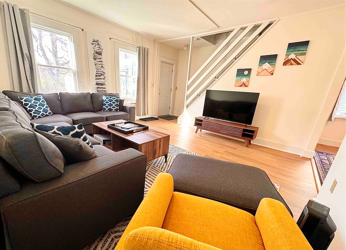 A cozy living area is presented, featuring a sectional sofa and an accent chair in vibrant yellow. A coffee table sits at the center, and a television is positioned against the wall. Sunlight filters through large windows, illuminating the warm-toned wooden floor.