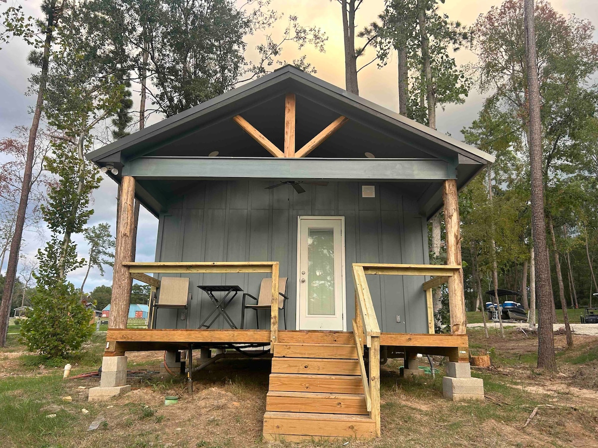A newly constructed cabin features a covered porch with wooden steps leading to a front door. The design includes exposed wooden beams and a combination of gray siding, creating a rustic appearance. Surrounding greenery complements the serene setting.