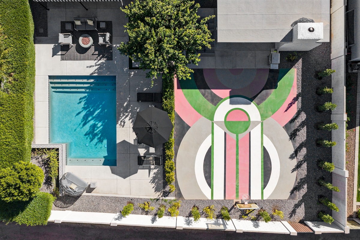 An aerial view captures a heated swimming pool surrounded by a spacious deck. A contemporary half-court basketball area is visible, showcasing vibrant geometric patterns. Lounge chairs and an umbrella provide shaded seating, while neatly arranged greenery enhances the outdoor space.