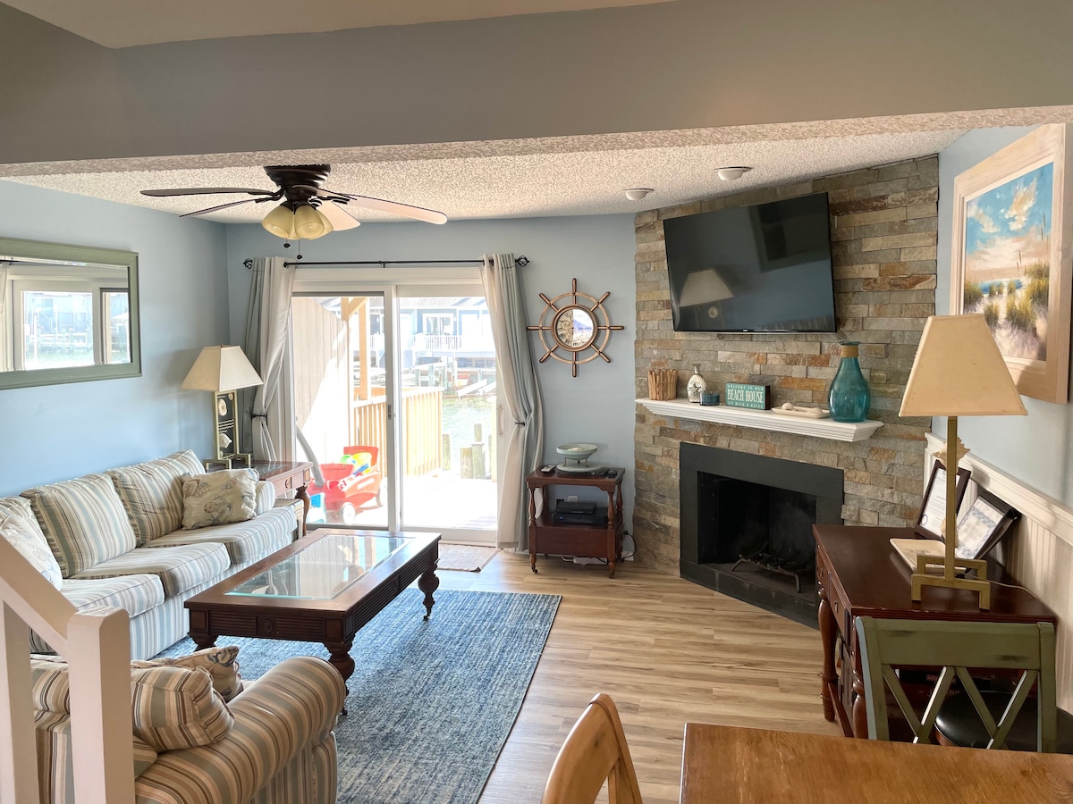 A comfortable living space is shown with a striped sofa and a glass coffee table. A flat-screen television is mounted above a stone fireplace. Natural light enters through sliding glass doors, leading to an outdoor area. Light blue walls complement the decor.
