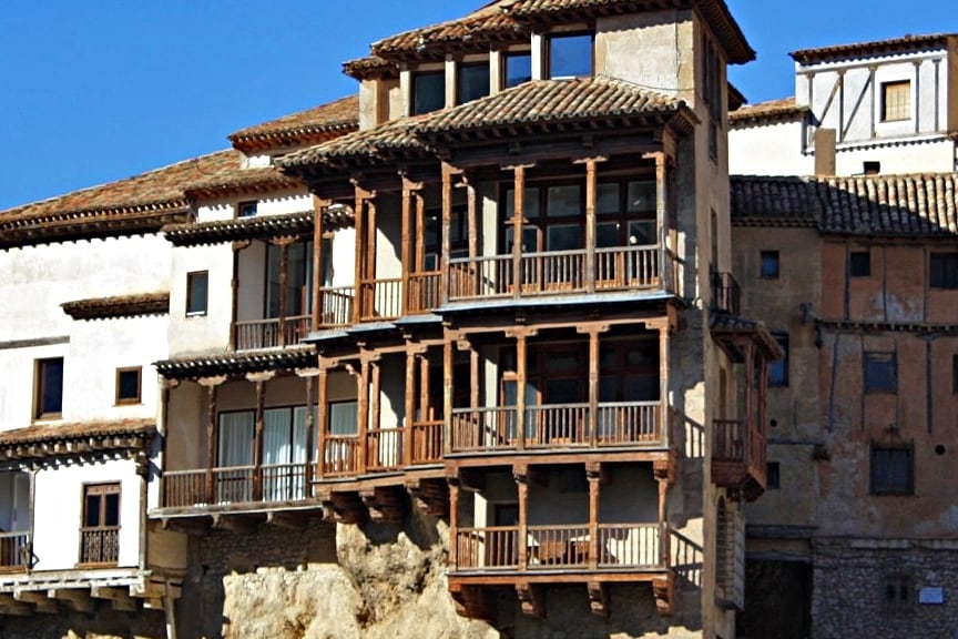 A multi-story building is featured with traditional wooden balconies and a stone foundation, showcasing a blend of rustic architecture and natural setting. The structure is situated on elevated land, surrounded by other buildings, with clear skies in the background.