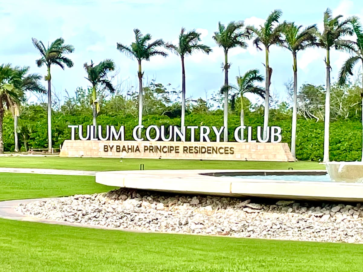The entrance sign of Tulum Country Club is displayed prominently, surrounded by lush green landscaping. Palm trees frame the area, adding to the tropical ambiance. The stone feature includes the text 'TULUM COUNTRY CLUB BY BAHIA PRINCIPE RESIDENCES,' welcoming guests to the community.