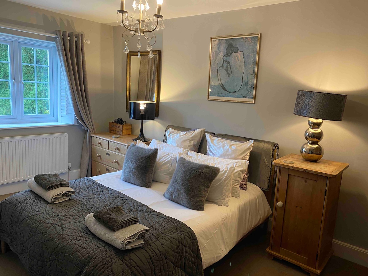 A well-appointed bedroom features a spacious bed adorned with plush pillows and a cozy quilt. Soft lighting is provided by a stylish lamp and chandelier, illuminating the room's neutral tones. A dresser and bedside cabinet offer functional storage, while a window allows natural light to enter.