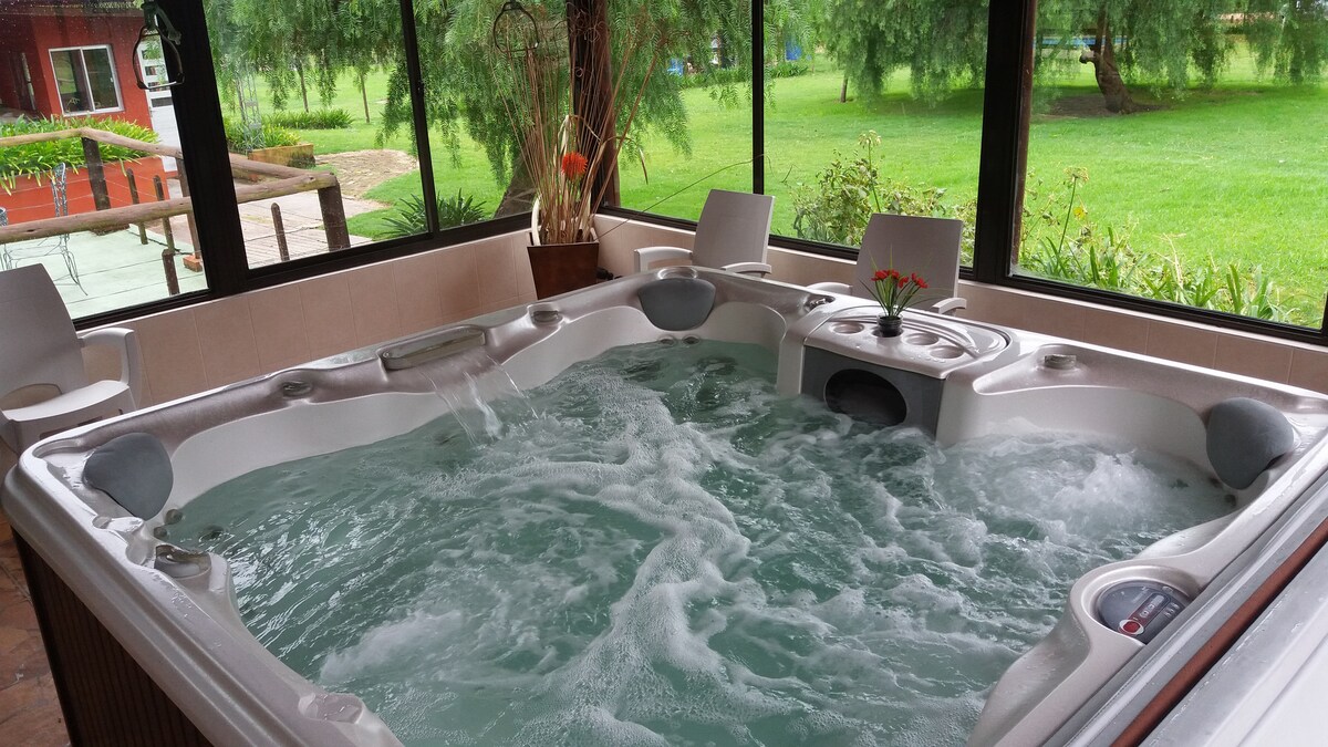 A spacious hot tub is situated in a sunlit room lined with large windows, allowing views of the lush greenery outside. Comfortable chairs are positioned nearby, enhancing the relaxation space. The bubbling water invites a tranquil atmosphere, suggesting a retreat from everyday life.