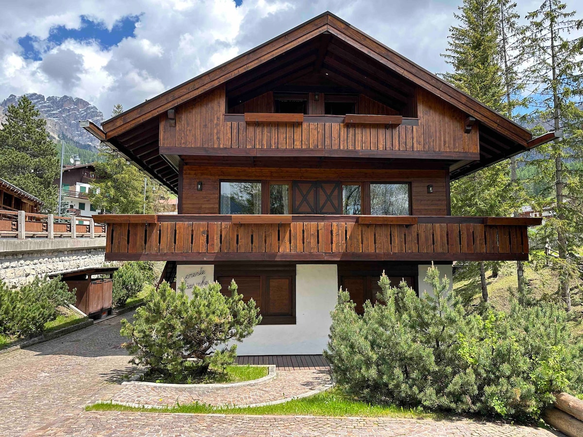 The exterior of a traditional mountain chalet is showcased, featuring wooden accents and a pitched roof. A landscaped area with shrubs surrounds the building, and large windows offer views into the interior. The structure is complemented by a scenic backdrop of greenery and mountains.