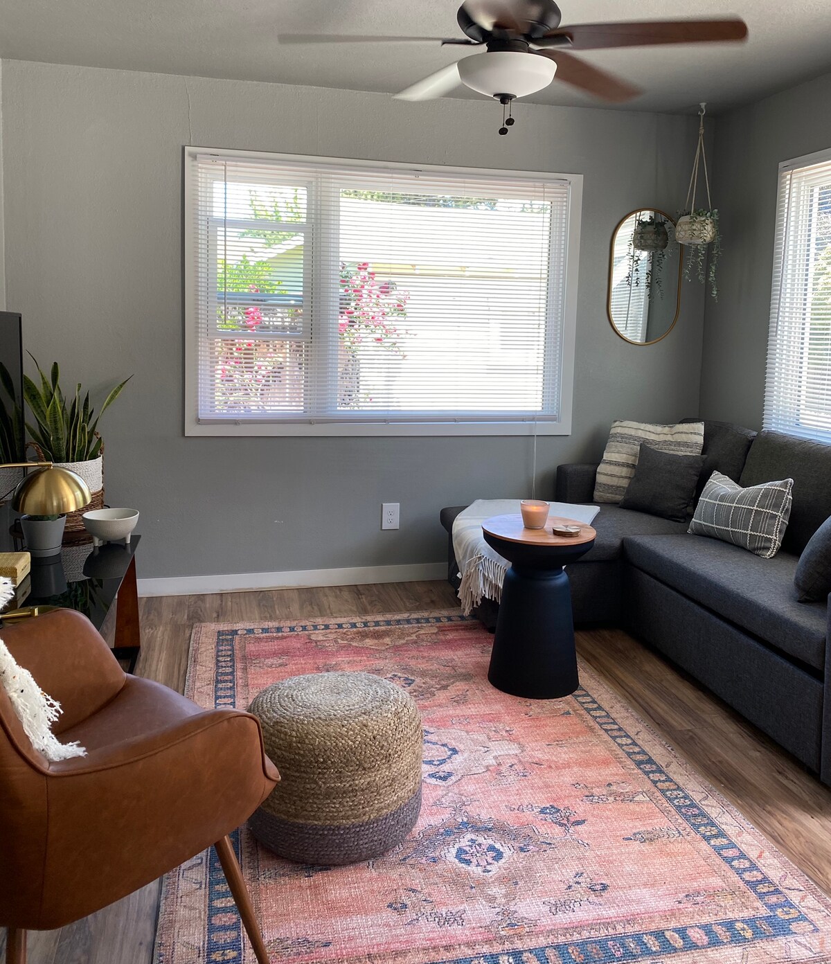 A cozy living room is presented, featuring a dark grey sofa, a small round black table, and a textured pouf. A woven rug with warm tones adds a decorative element. Natural light filters through large windows adorned with simple white blinds, highlighting a plant and wall decor.