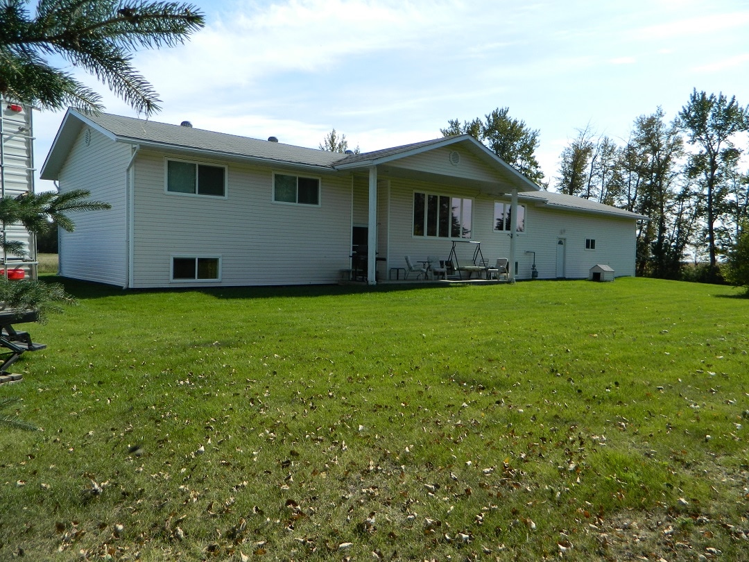 A white, single-story house is set on a spacious lawn, surrounded by trees. The back porch is visible, featuring a few chairs arranged for outdoor seating. The grassy area showcases scattered fallen leaves, creating a natural setting.