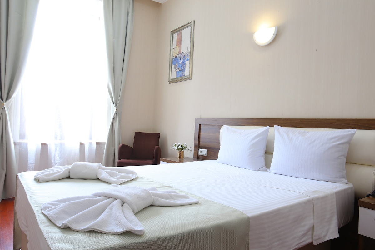 A guest room features a spacious bed covered with crisp linens and complemented by decorative towels arranged on top. Natural light filters through a large window, which is dressed with light curtains. A chair and decorative touch with flowers are also visible.