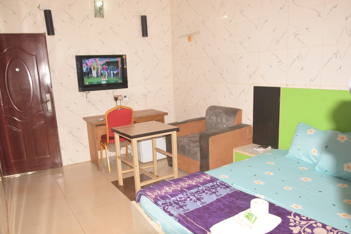 A compact living space features a double bed with a light blue cover and patterned pillows. A small table with two chairs is positioned nearby, alongside a comfortable couch. A television is mounted on the wall, providing entertainment options.