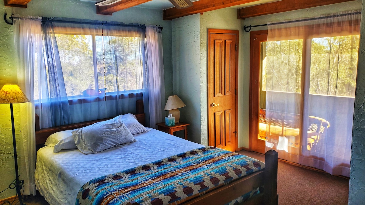 A cozy bedroom features a spacious bed with decorative pillows, soft lighting from a nearby lamp, and windows allowing natural light to fill the room. A private balcony with seating is accessible through glass doors, enhancing the views of the surrounding nature.