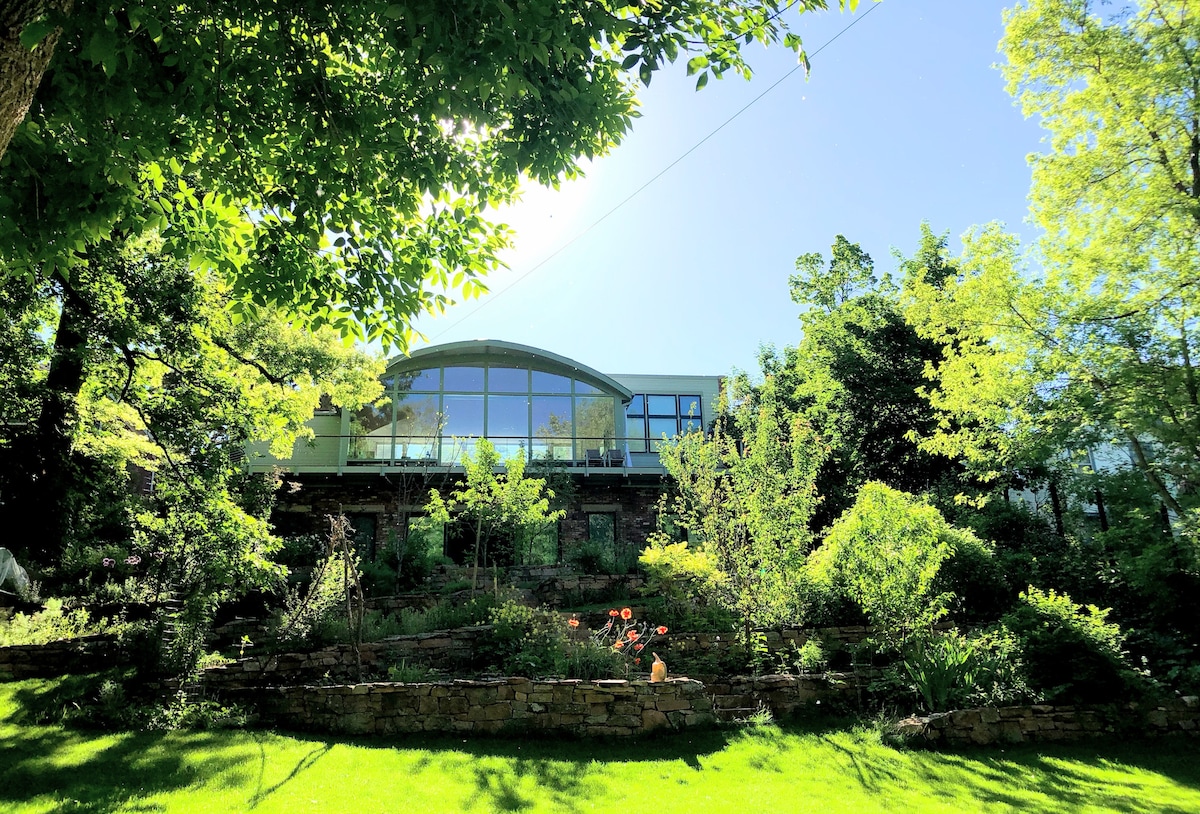A modern home is showcased, set on elevated ground with large glass windows reflecting the surrounding nature. Lush greenery and flowering plants create a vibrant garden, along with stone pathways leading through the landscape, enhancing the serene outdoor setting.