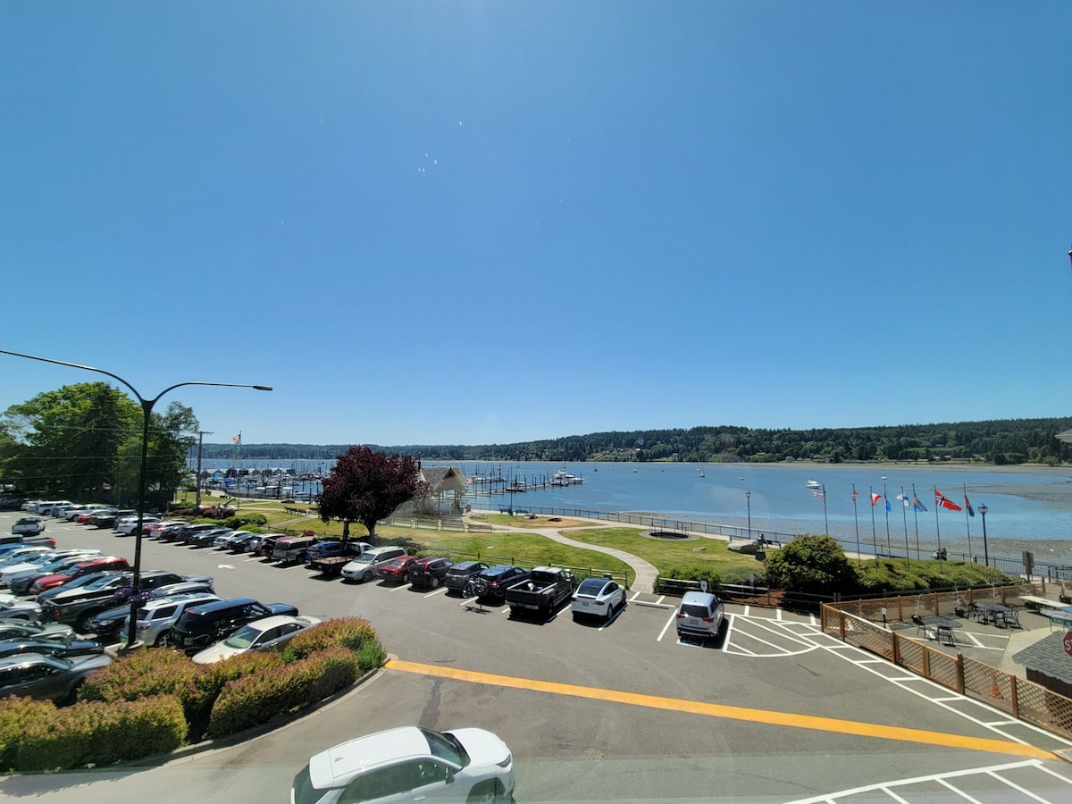 A view of the scenic Poulsbo waterfront is presented, featuring a marina dotted with boats. In the foreground, a parking area is visible, with several cars parked. The landscape is complemented by green trees and flags lining the shore, all under a clear blue sky.