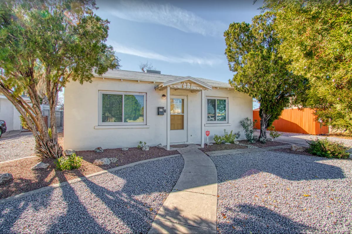 A single-story home features a simple entrance surrounded by well-maintained landscaping. A curved walkway leads to a front door with glass paneling, while gravel grounds and shrubs provide a neat foundation. Light-filtering trees offer shade, highlighting the home's inviting exterior.