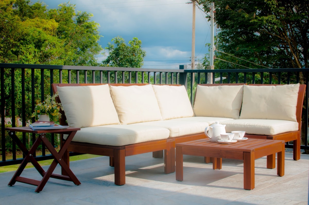 An outdoor seating area features a spacious sectional sofa with light-colored cushions and a wooden coffee table. A small side table is positioned beside the seating, complemented by a decorative flower arrangement. Lush greenery is visible in the background under a cloudy sky.