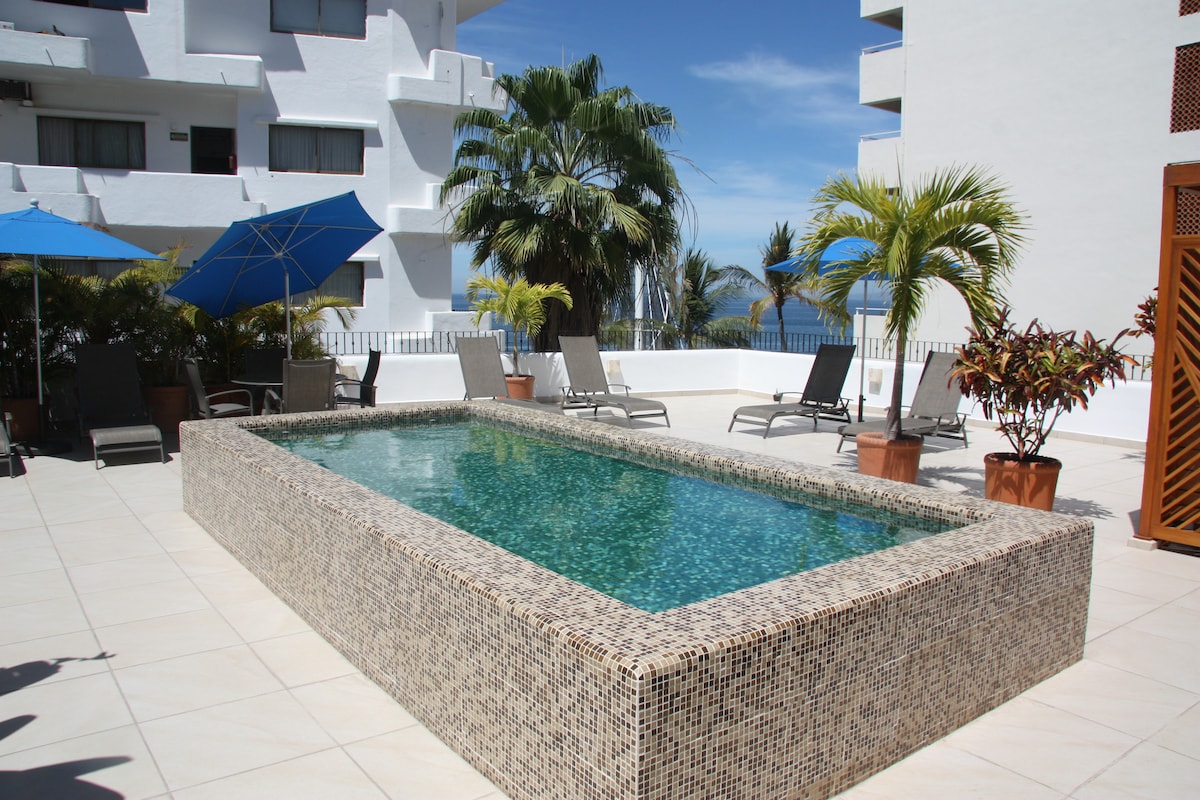A tranquil pool area is framed by lush palm trees and blue umbrellas. Lounge chairs provide relaxation space beside the water, with a modern tiled pool reflecting the sky’s colors. The surrounding area features white tiled flooring, enhancing the serene ambiance.