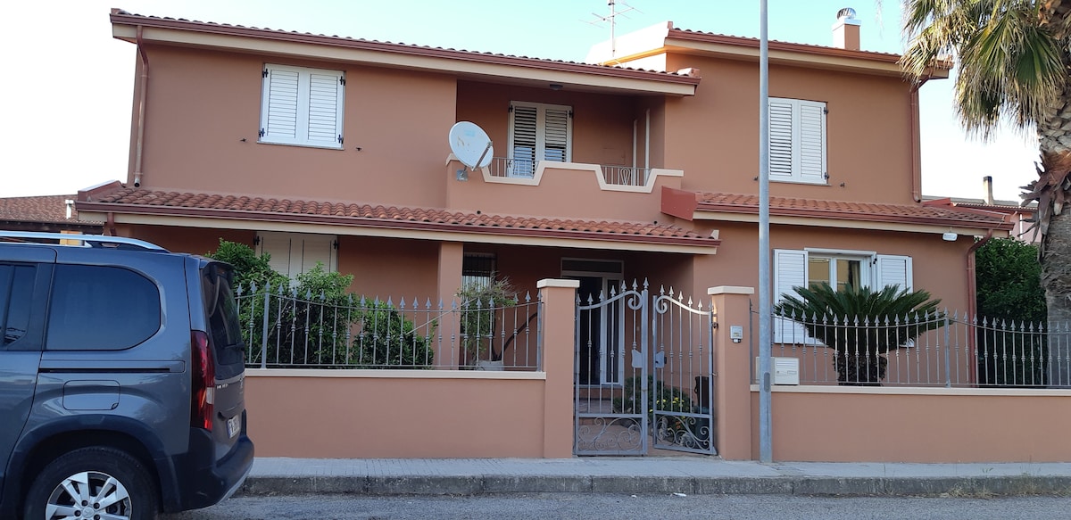 The exterior of a two-story house is showcased with a warm orange facade and a terracotta roof. A metal gate leads to a well-kept garden with palm trees. Windows with white shutters are visible, providing a welcoming appearance to the entryway.