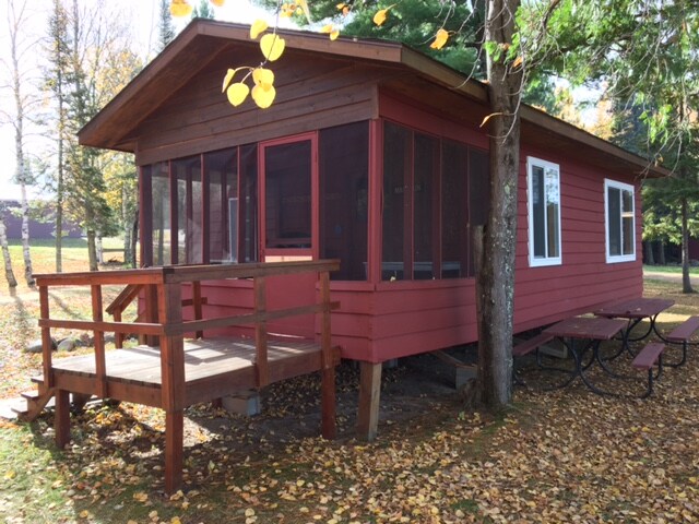 A cabin exterior is shown, featuring a red wooden structure with a screened-in porch. A wooden deck leads to the entrance, surrounded by vibrant fallen leaves. A picnic table sits under a tree nearby, enhancing the outdoor appeal of the space.