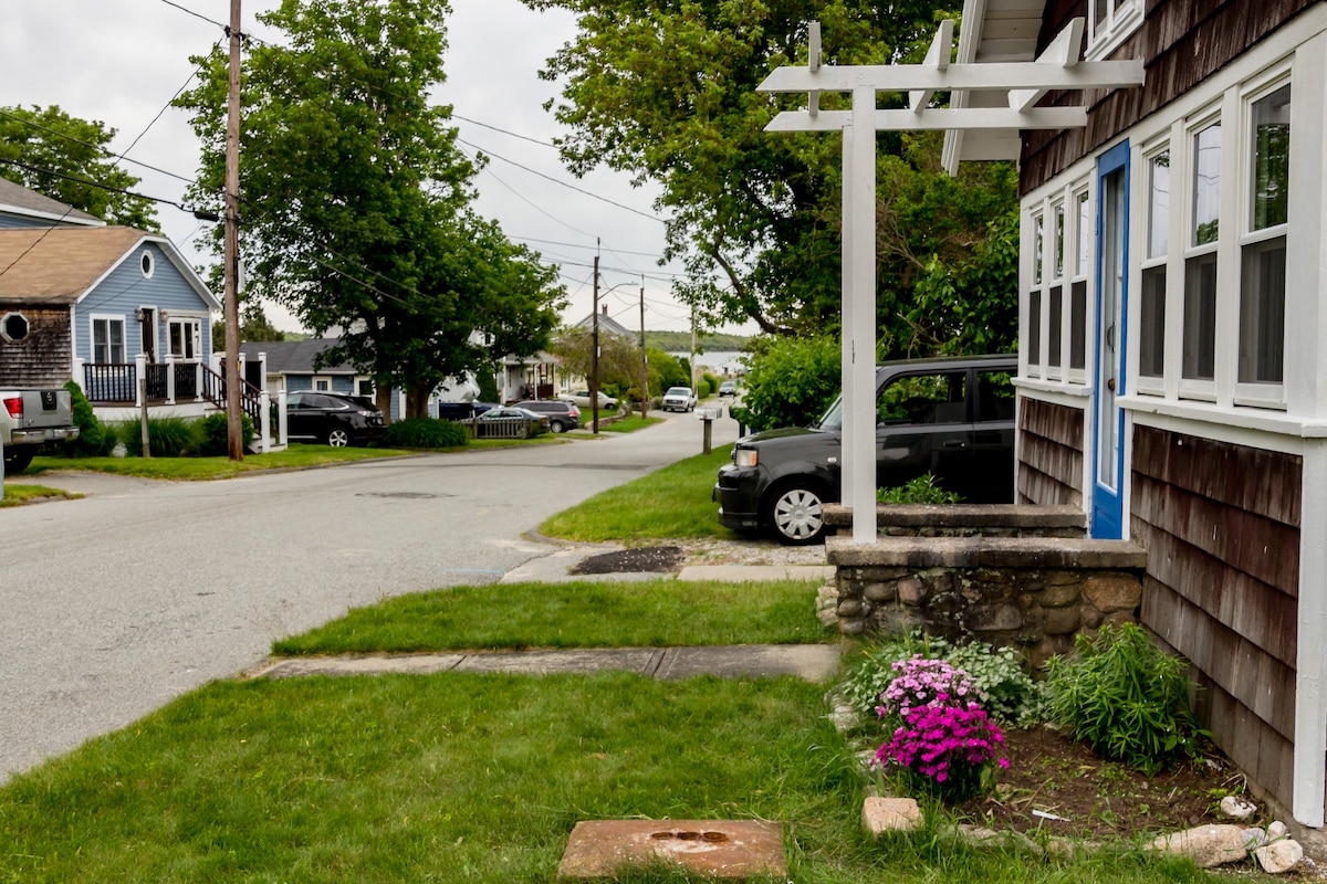 The exterior of the cottage is shown, featuring a welcoming blue door and a well-maintained front yard with colorful flowers. The view includes a quiet street lined with trees and nearby houses, providing a glimpse of the neighborhood atmosphere.