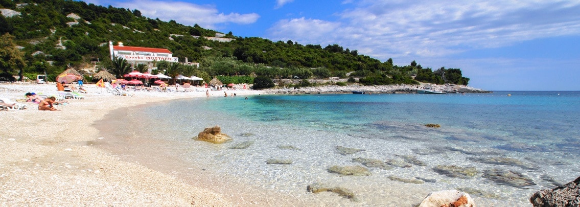 A serene beach scene captures clear turquoise waters lapping against a sandy shore, with gentle waves revealing smooth pebbles beneath the surface. Sunbathers can be seen enjoying the sun under umbrellas, with lush greenery framing the beach area in the background.