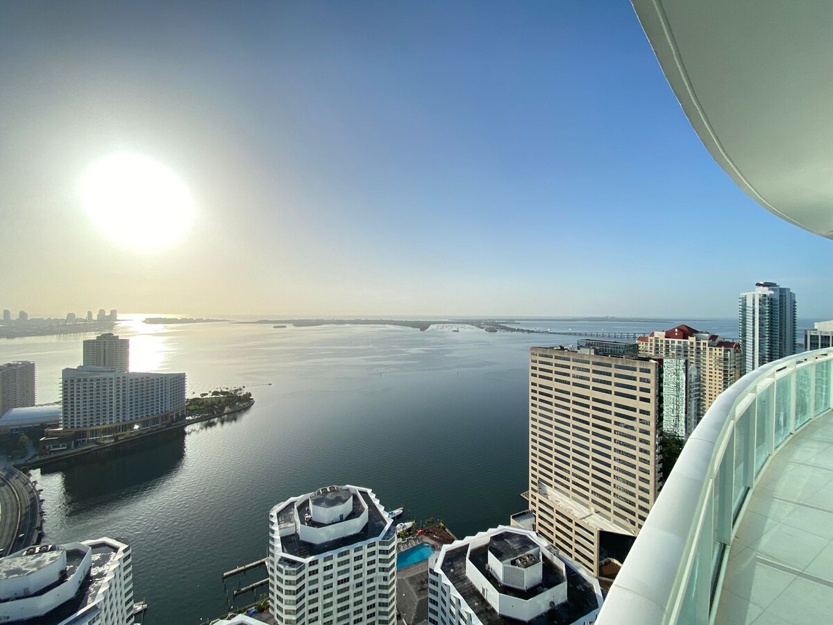 Bright, clear views of the surrounding waterfront and city skyline are presented from a spacious balcony. The sun casts a warm glow over the horizon, reflecting on the water. Architectural details of nearby buildings are visible below.