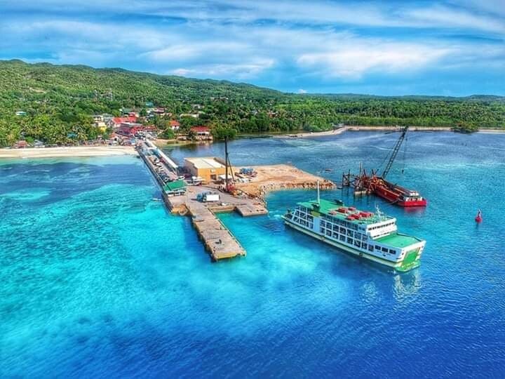 An aerial view captures a dock beside vibrant turquoise waters. The dock features large vessels and construction equipment, while the shoreline is lined with greenery. Nearby, a small settlement with buildings is visible, nestled among rolling hills and trees.